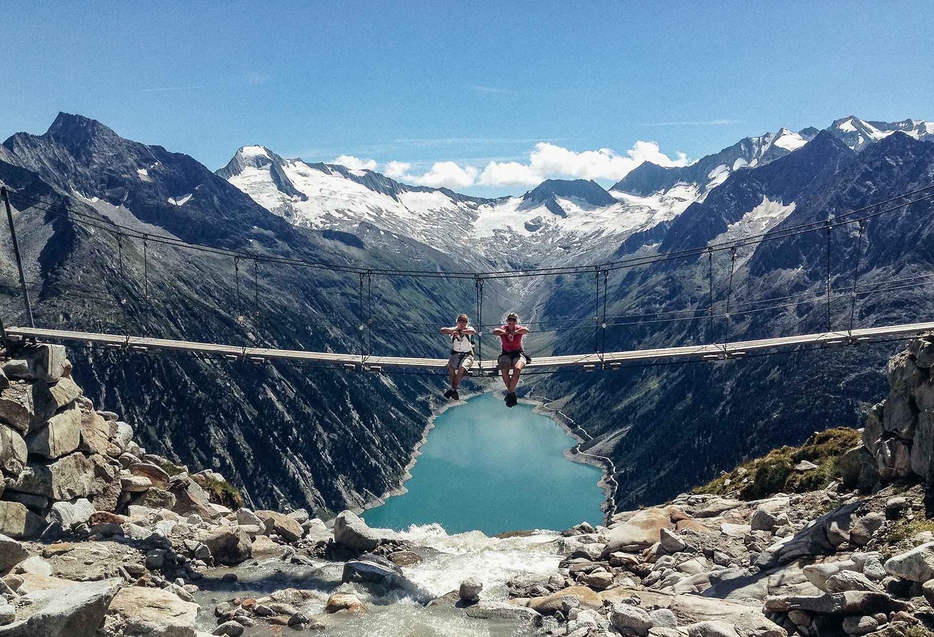 Zwei Wanderer sitzen auf einer Hängebrücke hoch über einem Bergsee